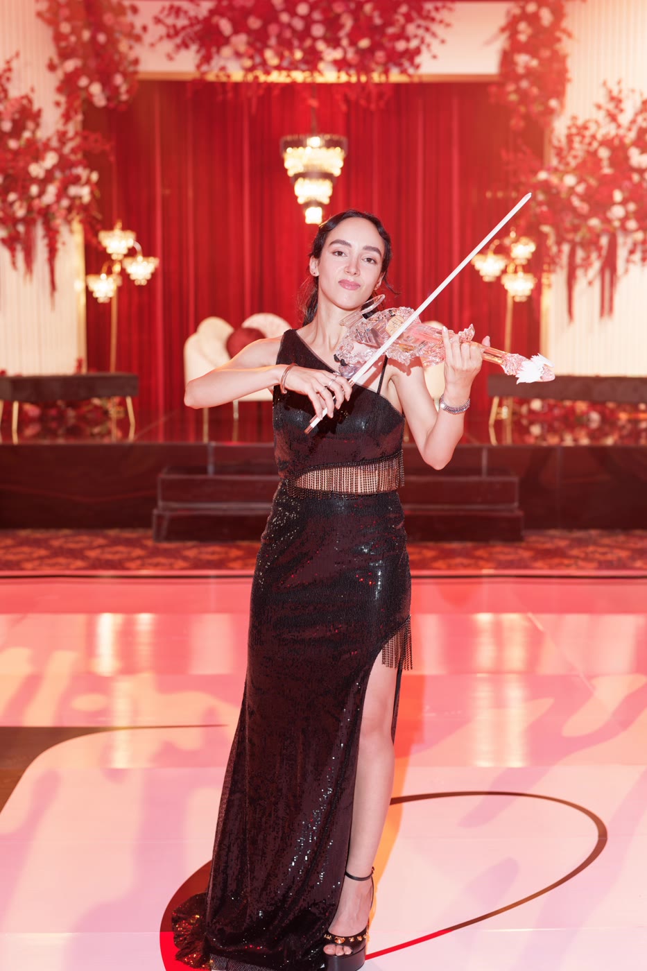 The transparent violin under Sadie’s bow on a red ballroom stage, its custom body catching the light
