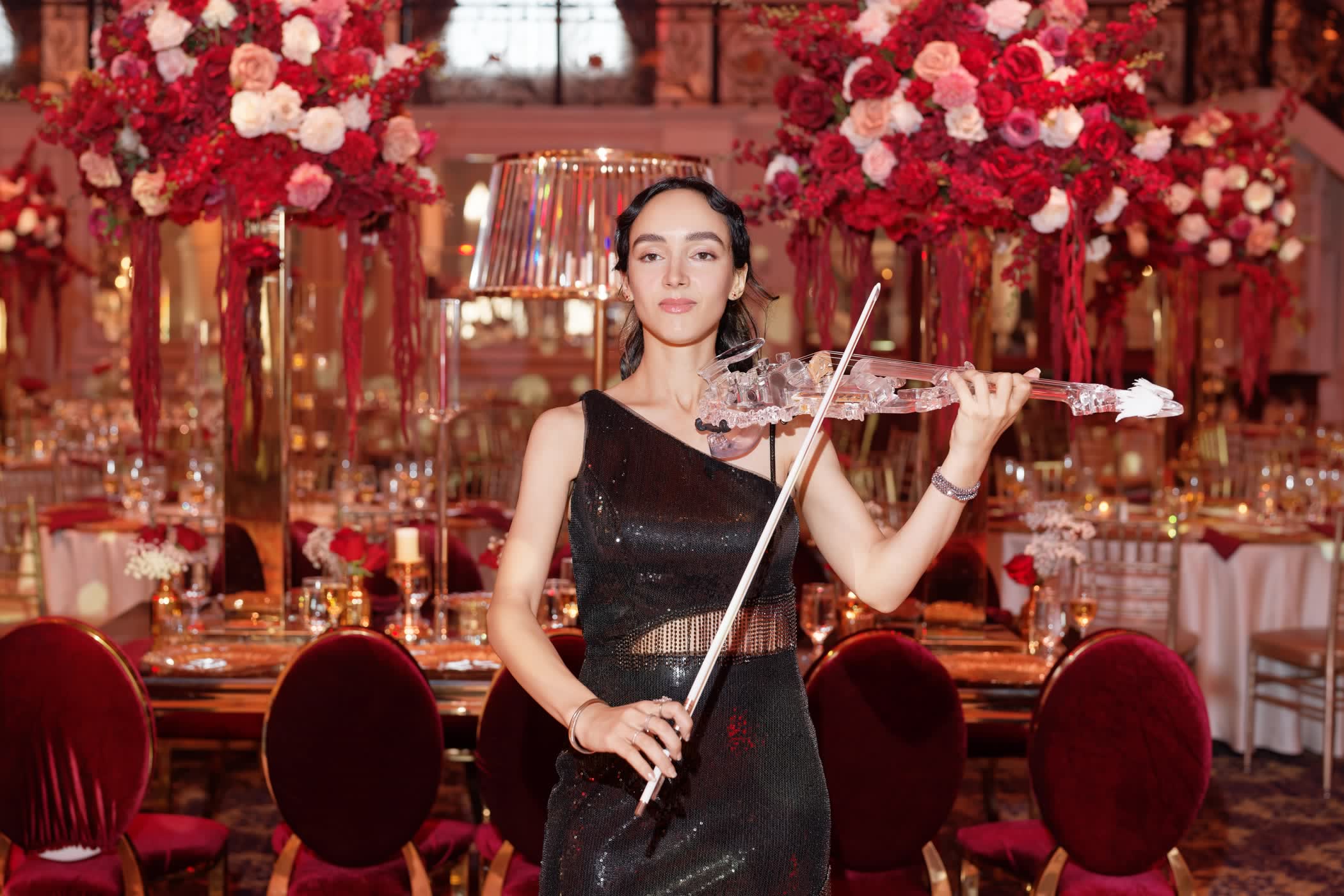 Sadie holding the transparent violin under a crystal lampshade centerpiece, red roses around her